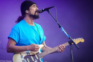 Musician wearing a blue shirt and beanie sings into a microphone while playing a sticker-covered electric guitar on stage with a purple backdrop.