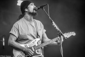 Male guitarist in a beanie sings into a microphone while playing a white Fender Stratocaster on stage, stickers visible on the guitar (black and white image).