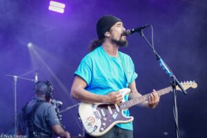 Male guitarist with beard and beanie singing into a microphone while playing a white Stratocaster on a purple-lit stage with stickers on the guitar surface.