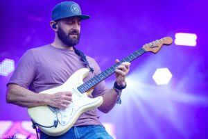 Male guitarist on stage playing a white Stratocaster, wearing a teal cap and a brown shirt, with purple stage lights in the background