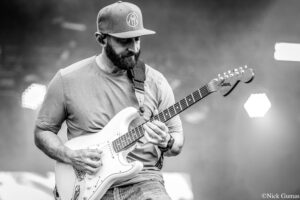 Black-and-white photo of a bearded guitarist wearing a cap, playing a white Fender Stratocaster on stage.