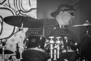 Drummer wearing a backwards cap and sunglasses performing on stage behind a drum kit.