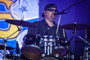 Drummer wearing sunglasses and a cap performs behind a drum kit on stage with microphones nearby.