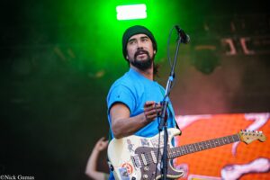 Male guitarist on stage wearing a blue shirt and black beanie, playing a sticker-covered Fender guitar with microphones nearby, green stage lights in background