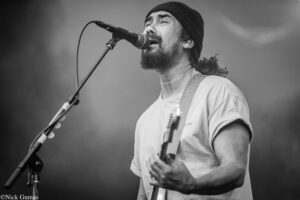 Male singer with a beard and beanie, singing into a microphone on stage with a guitar strap visible