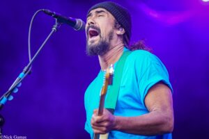 Male musician in a blue shirt and black beanie singing into a microphone on stage with purple lighting in the background.