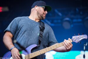 Male bass guitarist performing on stage, wearing a dark cap and sunglasses, close-up of purple bass guitar neck and fretboard in hand