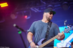 Male bass guitarist on stage wearing sunglasses and a dark cap, playing a purple bass guitar amid red/purple stage lighting.