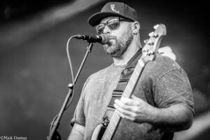 Male guitarist and vocalist wearing a cap and sunglasses, singing into a microphone on stage while playing an electric guitar in black-and-white.