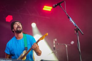 Guitarist in a blue shirt and black beanie plays a cream-colored electric guitar on a stage with red lighting behind him.