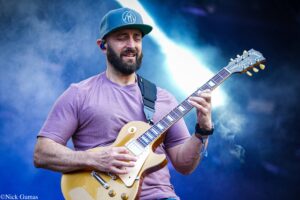 Male guitarist in a purple shirt and cap, playing a gold Les Paul–style electric guitar on a blue-lit stage with smoke.