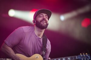 Male guitarist singing and playing a sunburst electric guitar on stage, wearing a cap and pink shirt under pink-purple lights.