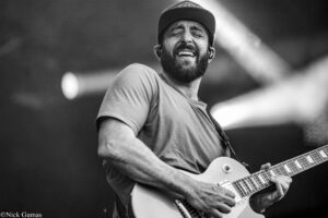 Black-and-white image of a smiling guitarist wearing a cap, playing an electric guitar on stage.