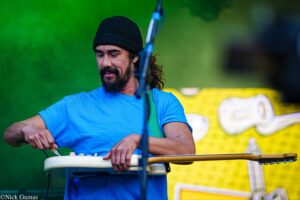 Bearded man in a blue shirt and black knit cap tunes a small keyboard outdoors on a stand.