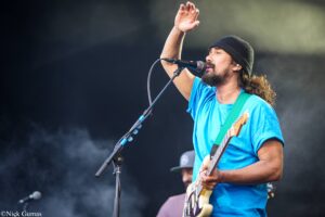 Male guitarist and vocalist wearing a blue shirt and black beanie performing on stage, raising his hand, singing into a microphone.