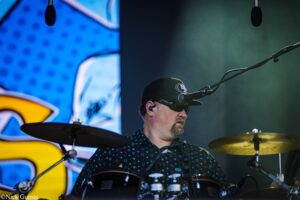 Drummer wearing a black cap and sunglasses, playing a drum kit on stage with a large blue screen behind him.