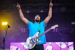 Guitarist on stage with arms raised, wearing a blue shirt and black beanie, playing a sticker-covered cream Fender Stratocaster.