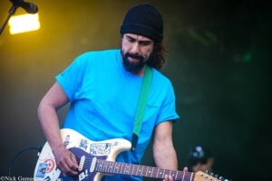 Male guitarist wearing a blue t-shirt and black beanie, playing a cream-colored guitar with stickers on an outdoor stage.