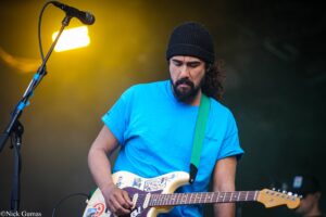 Male guitarist on stage wearing a bright blue t-shirt and black beanie, playing a white sticker-covered electric guitar with a microphone nearby.