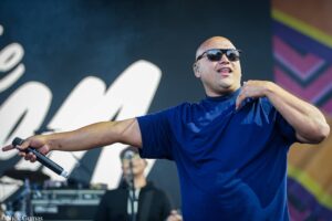 Male singer on stage, wearing sunglasses and a blue t-shirt, arms outstretched while holding a microphone.