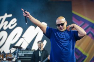 Male performer in a blue shirt and sunglasses raises a microphone on stage during a live concert, with a band and colorful backdrop in the background.