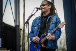 Guitarist wearing a blue denim jacket and black hoodie, sunglasses, singing into a microphone on stage with a blue electric guitar.