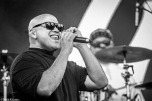 Male singer wearing sunglasses, holding a microphone and performing on stage; drummer in the blurry background.