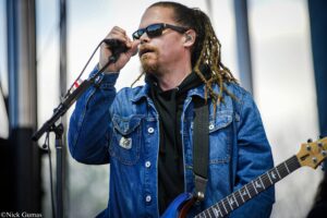 Male musician singing into a microphone on stage, wearing a denim jacket and sunglasses with dreadlocks visible.