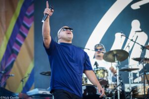 Male vocalist on stage raises microphone toward the crowd, wearing a blue shirt and sunglasses, with a drummer in the background.