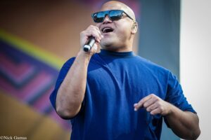 Male singer in a blue shirt wearing sunglasses, singing into a handheld microphone on stage on a sunny day.”