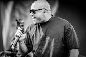Black-and-white photo of a bald man wearing sunglasses, singing into a handheld microphone on stage.