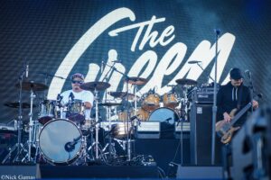 Band performing on an outdoor stage with a large white cursive backdrop; drummer center-left, guitarist right, drums and amps visible.