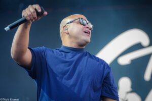 Male singer wearing sunglasses and a blue shirt performs on stage, holding a microphone far from the mouth, with a dark backdrop behind him.