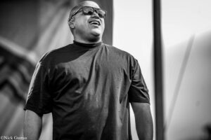 Black-and-white photo of a bald man on stage, wearing sunglasses and an in-ear monitor, smiling while performing.