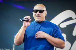 Male performer wearing sunglasses and a blue shirt, singing into a handheld microphone on stage.
