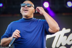Male performer singing into a microphone on stage, wearing sunglasses and a blue shirt, with purple stage lights in the background.