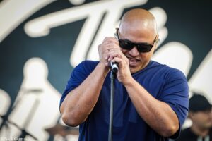 Male singer wearing sunglasses and a blue shirt, gripping a microphone on stage.