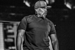Male performer wearing a cap and black T-shirt on stage, looking to the right with a serious expression.