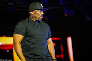 Male performer on stage in a black t-shirt and cap, looking to the side amid colorful stage lights underdark background.