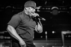 Male singer on stage wearing a baseball cap, gripping a microphone and performing passionately.