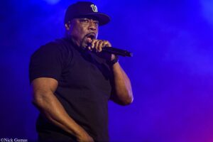 Male singer in a black t-shirt and cap holding a microphone on a blue-lit stage during a live performance.