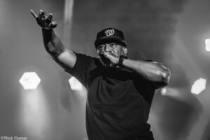 Male performer on stage wearing a cap with a W logo, singing into a microphone and reaching toward the audience with his left arm raised.