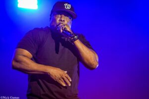 Male singer on stage, wearing a black t-shirt and cap, singing into a microphone under blue stage lights.