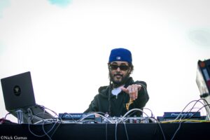 DJ wearing a blue cap and sunglasses performs at a mixing console with a laptop and various cables visible onstage.