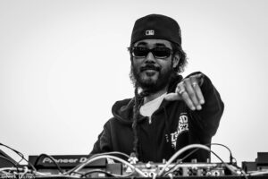 Black-and-white shot of a DJ wearing a cap and sunglasses, reaching toward a cluttered mixing console with cables.