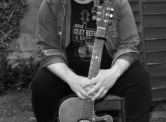 Man wearing a cowboy hat sits outdoors with an acoustic guitar in his lap, black-and-white photo beside a shed and dirt yard.