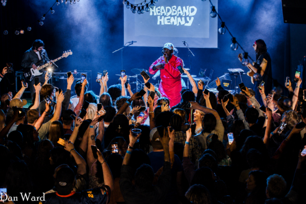 Crowd at a live concert raises phones toward a performer in a bright pink jumpsuit on a blue-lit stage with a screen reading Headband Henny in the background.