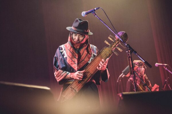 Musician in a hat and red scarf playing a long-necked string instrument on a dimly lit stage, with microphones nearby.