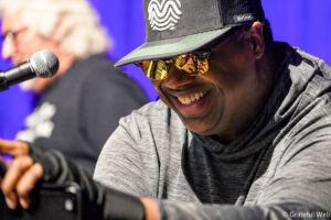 Smiling performer wearing a black cap with a white logo and gold mirrored sunglasses, singing into a microphone on stage.