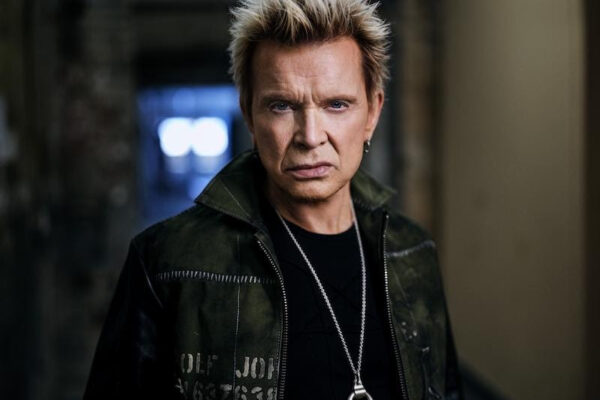 Serious-looking man with spiky blonde hair in a dark jacket and a silver pendant, standing in a dim hallway.